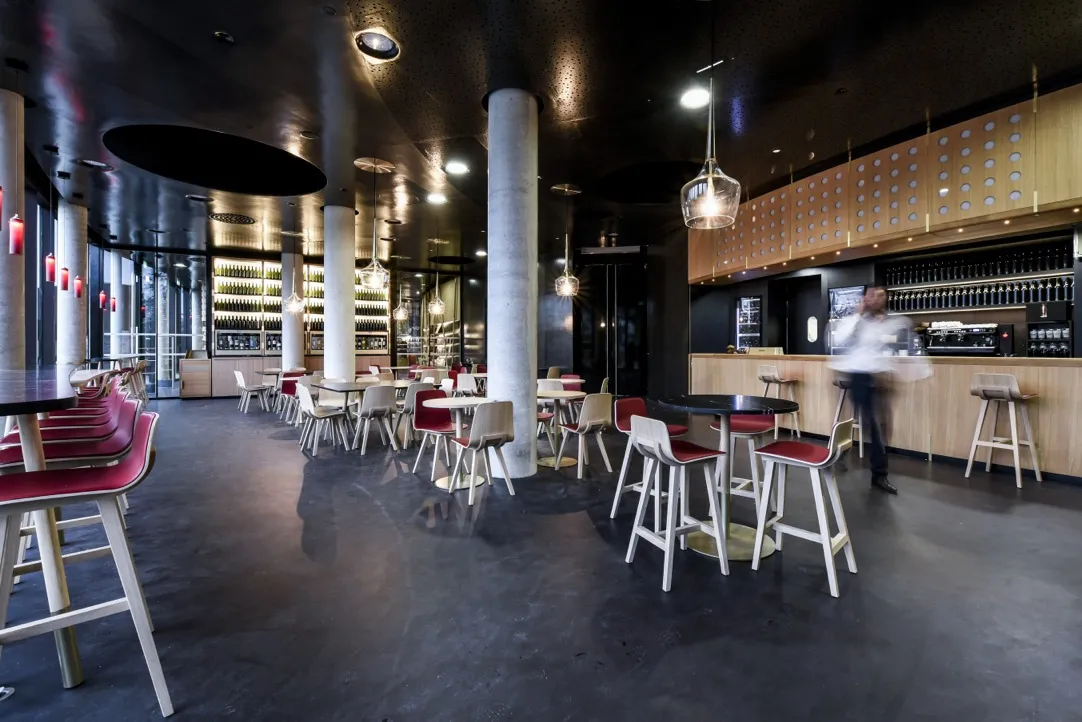 Modern interior of Latitude 20 wine bar in Bordeaux featuring high tables, red and white chairs, light wood bar counter, white columns, contemporary lighting, and staff member in background.