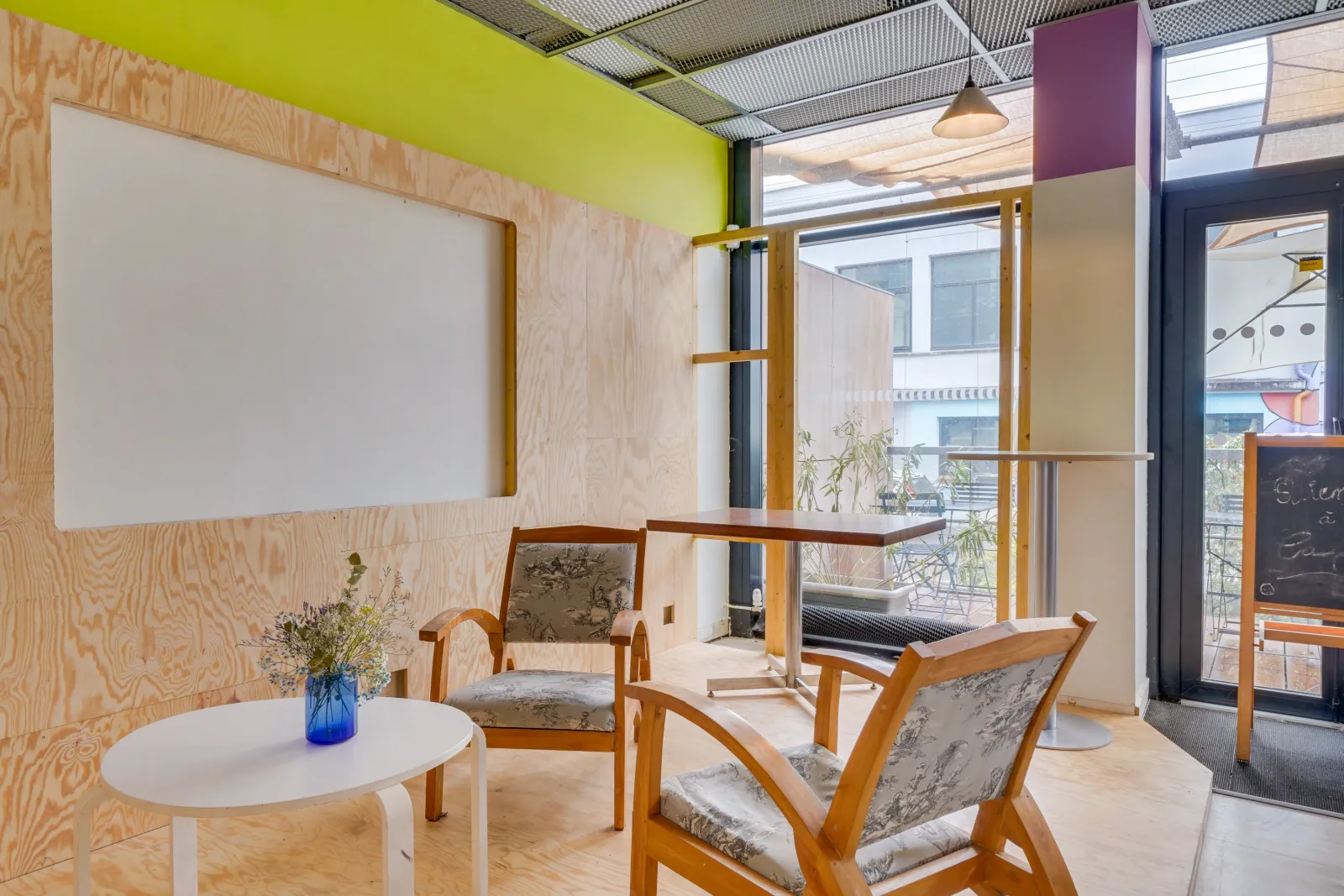 Modern relaxation area featuring two wooden armchairs with toile de Jouy cushions, a small white round table with a blue vase of flowers, plywood-clad walls, a blank poster, and glass doors opening onto a balcony with views of Bordeaux.