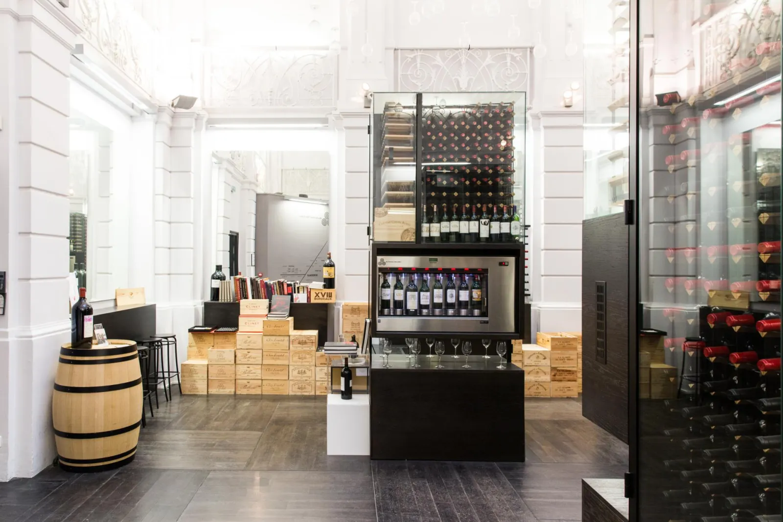 Max Bordeaux Wine Gallery showing a tasting bar with glass wine cellar display, Bordeaux wine bottles, wooden barrels, storage crates and modern furnishings in a sleek white space.