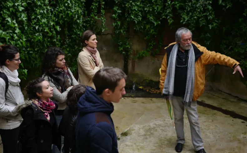 A tour guide in a yellow jacket explains the history of the neighborhood to a group of visitors in an alley of Old Lormont in Bordeaux, surrounded by ivy.
