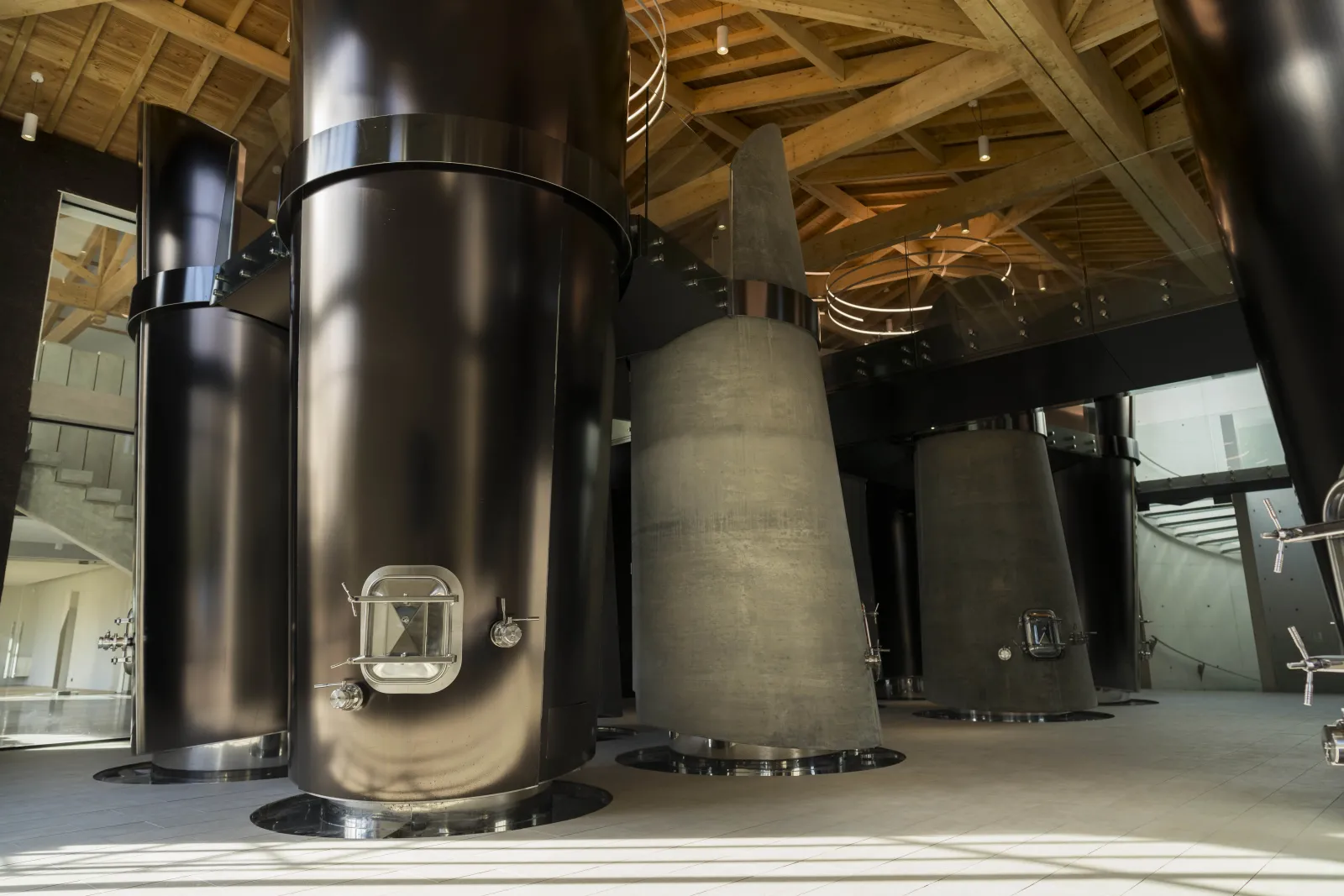 Stainless steel and concrete fermentation tanks in a Bordeaux winery cellar at Château Montlabert, beneath an exposed wooden structure.