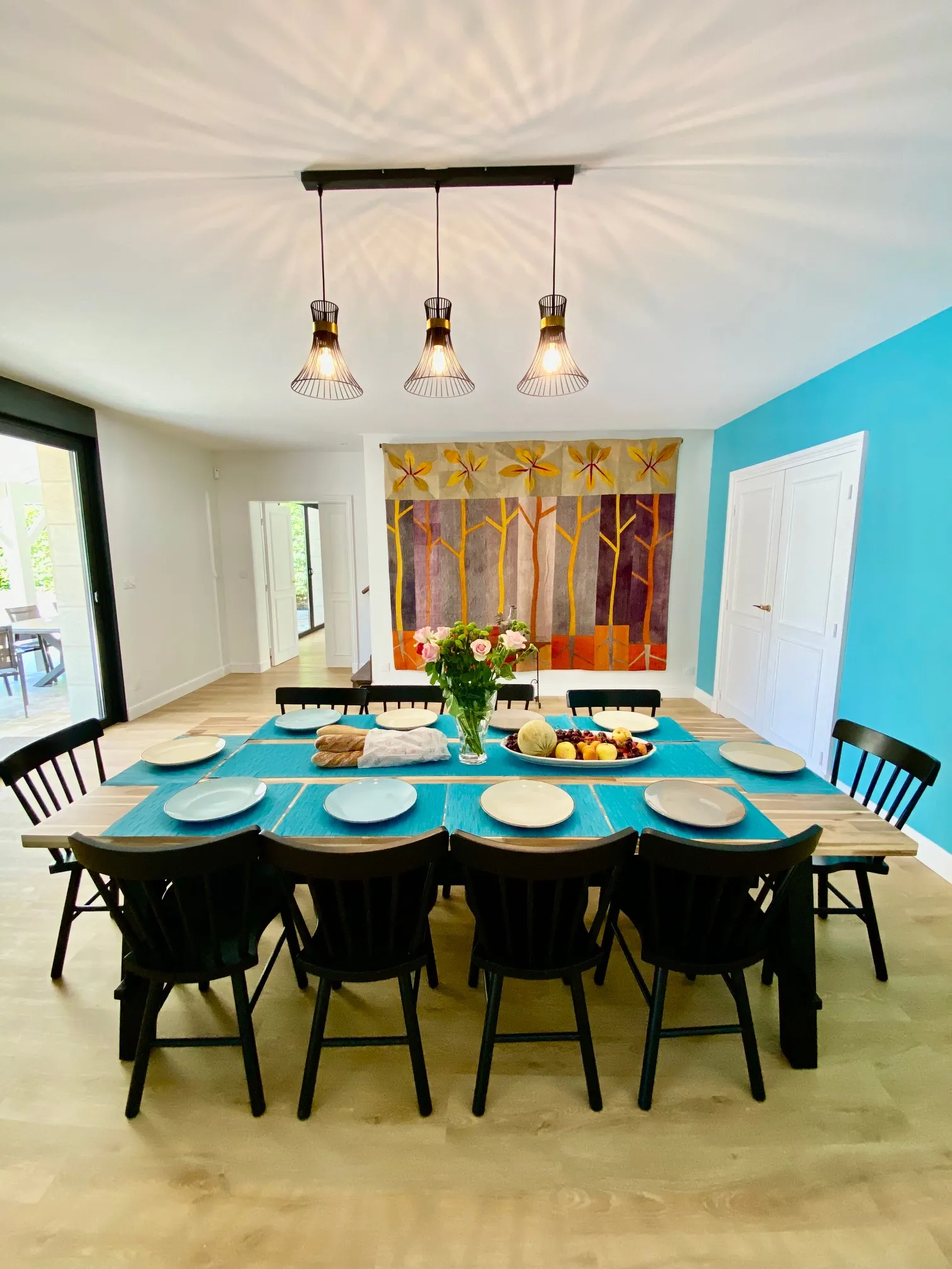 Modern dining room with black table, matching chairs, turquoise tablecloths, brass pendant lights, turquoise and white walls, abstract wall art, and floral decorative elements at La Maison L'Eperon in Bordeaux.