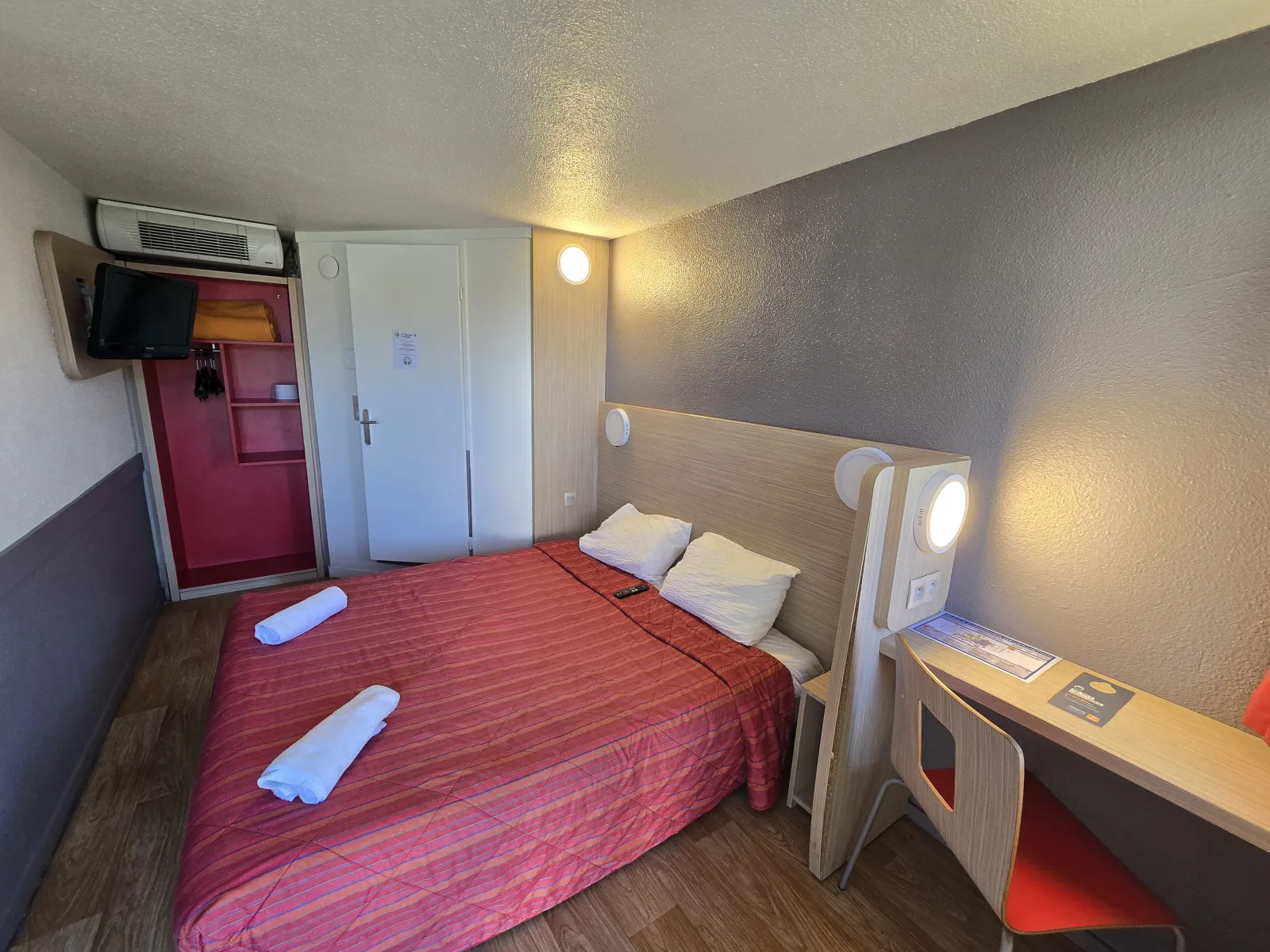 Modern hotel room featuring a double bed with red linens, two white pillows, desk with red chair, wall-mounted air conditioning, flat-screen TV, and ambient LED lighting.