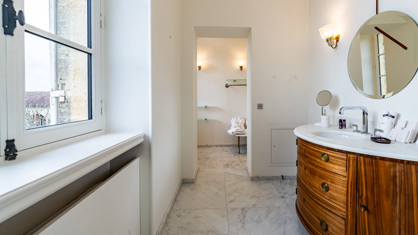Elegant bathroom at Château Malescasse in Bordeaux featuring a round white sink, mahogany vanity furniture, circular mirrors, gray and white marble flooring, bright window, and access to an adjacent bedroom.
