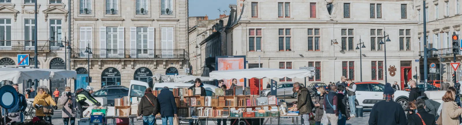 Outdoor market on Quai des Chartrons in Bordeaux featuring second-hand book vendors under white umbrellas, with Haussmann-style buildings in the background and shoppers browsing the stalls.