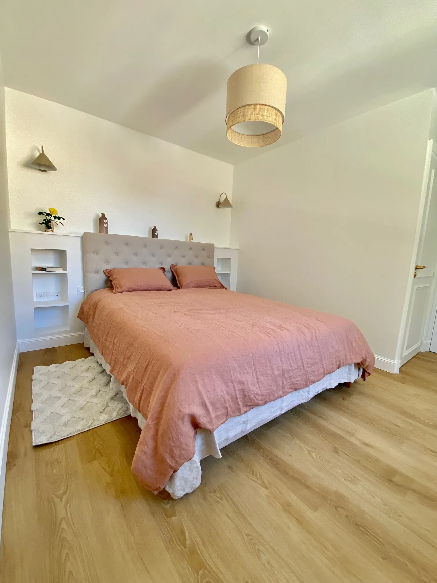 Modern bedroom featuring a queen-size bed with pink linens, upholstered gray headboard, white built-in storage, wooden pendant light, light wood flooring, and minimalist decor.