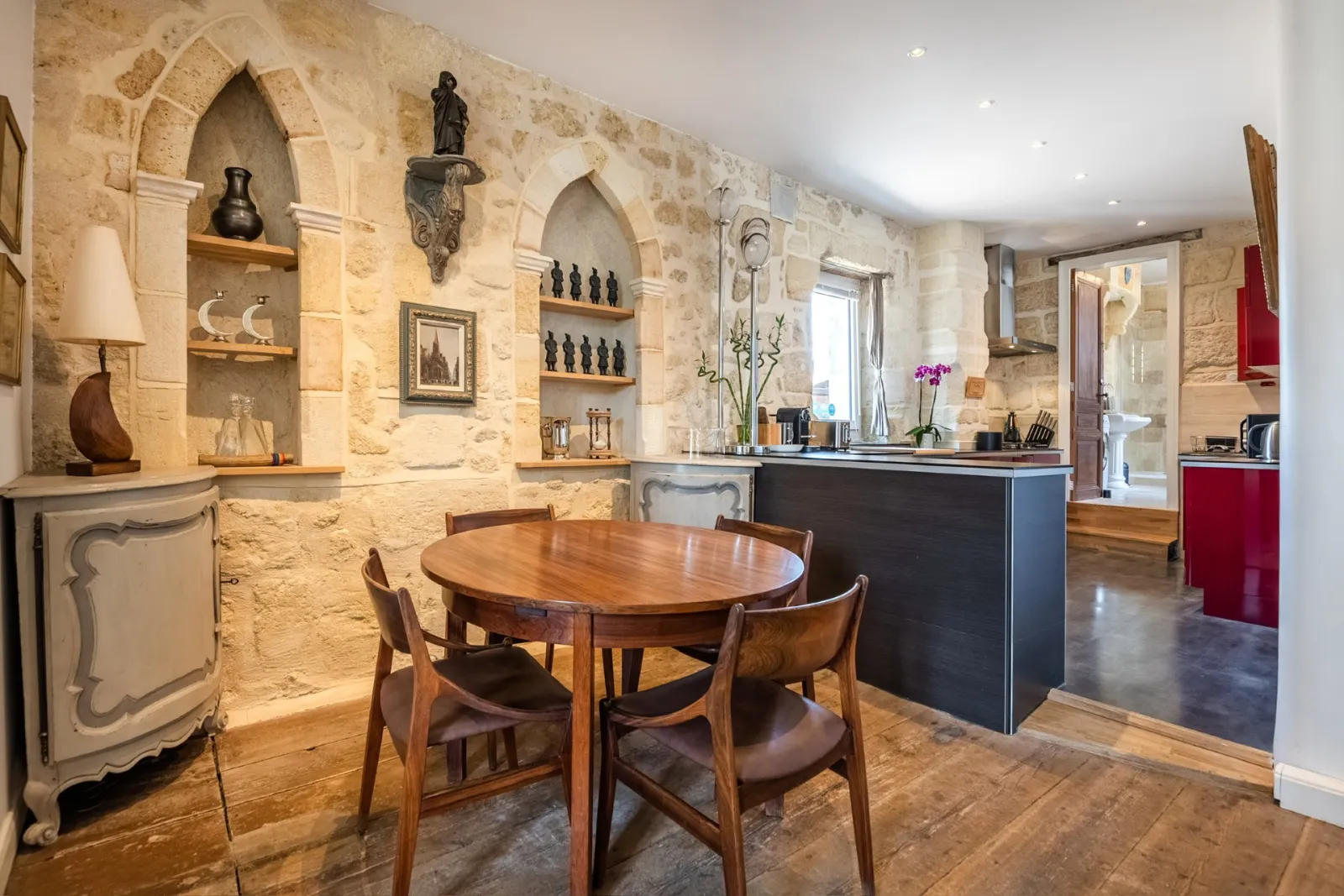 Elegant kitchen-dining room with cut stone walls, round wooden table, black central counter, decorative niches and statues, at La Monnoye in Bordeaux