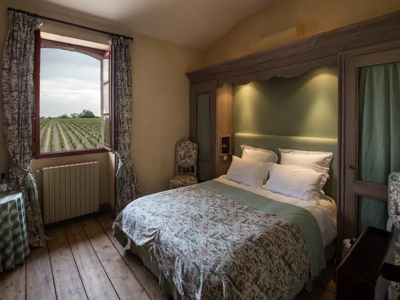 Elegant guest bedroom at Château Giscours featuring a king-size bed, dark wood furniture, window overlooking Bordeaux vineyards, patterned curtains, vintage radiator, and classic décor.