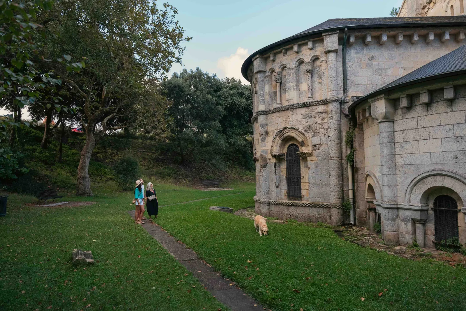 Two visitors and a dog in front of the Basilica of Notre-Dame-de-la-Fin-des-Terres in Bordeaux, a Romanesque building with cylindrical towers, surrounded by lawn and greenery.