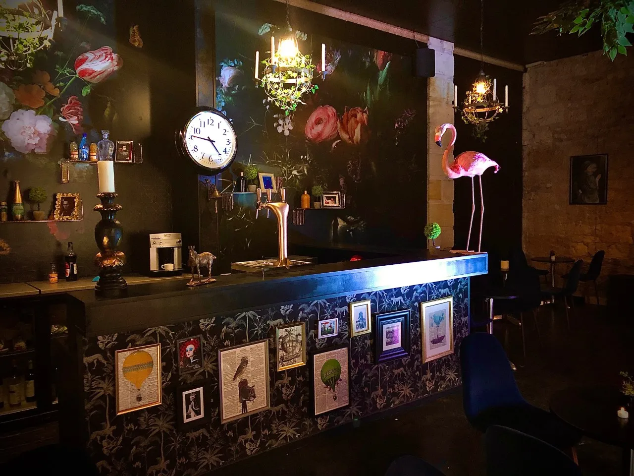 Elegant bar at the Théâtre des Chartrons in Bordeaux featuring an illuminated blue counter, black walls with painted flowers, clock, neon pink flamingo, golden chandeliers, and framed artwork decorating the counter