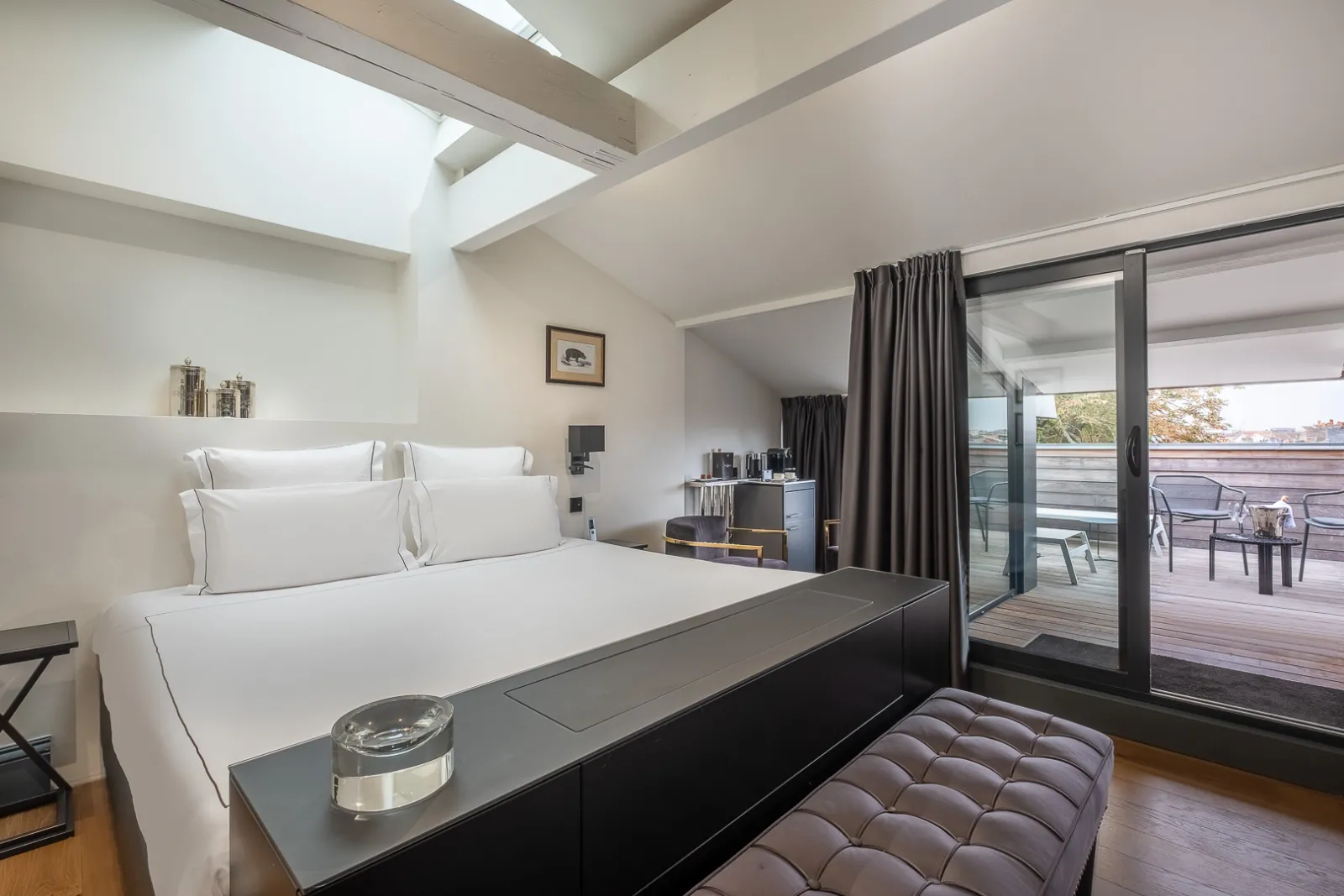 Modern bedroom at Palais Gallien Hôtel & Spa in Bordeaux featuring a king-size bed with white linens, exposed beam ceiling, panoramic glass doors opening onto a terrace, and contemporary black and gray furnishings.