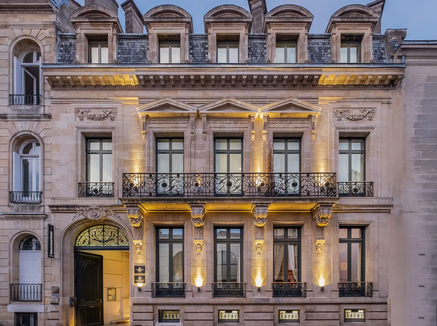 Illuminated facade of Palais Gallien Hôtel & Spa in Bordeaux, Haussmann-style building featuring three stories of windows with gilt moldings, wrought iron balconies and ornamental stonework.