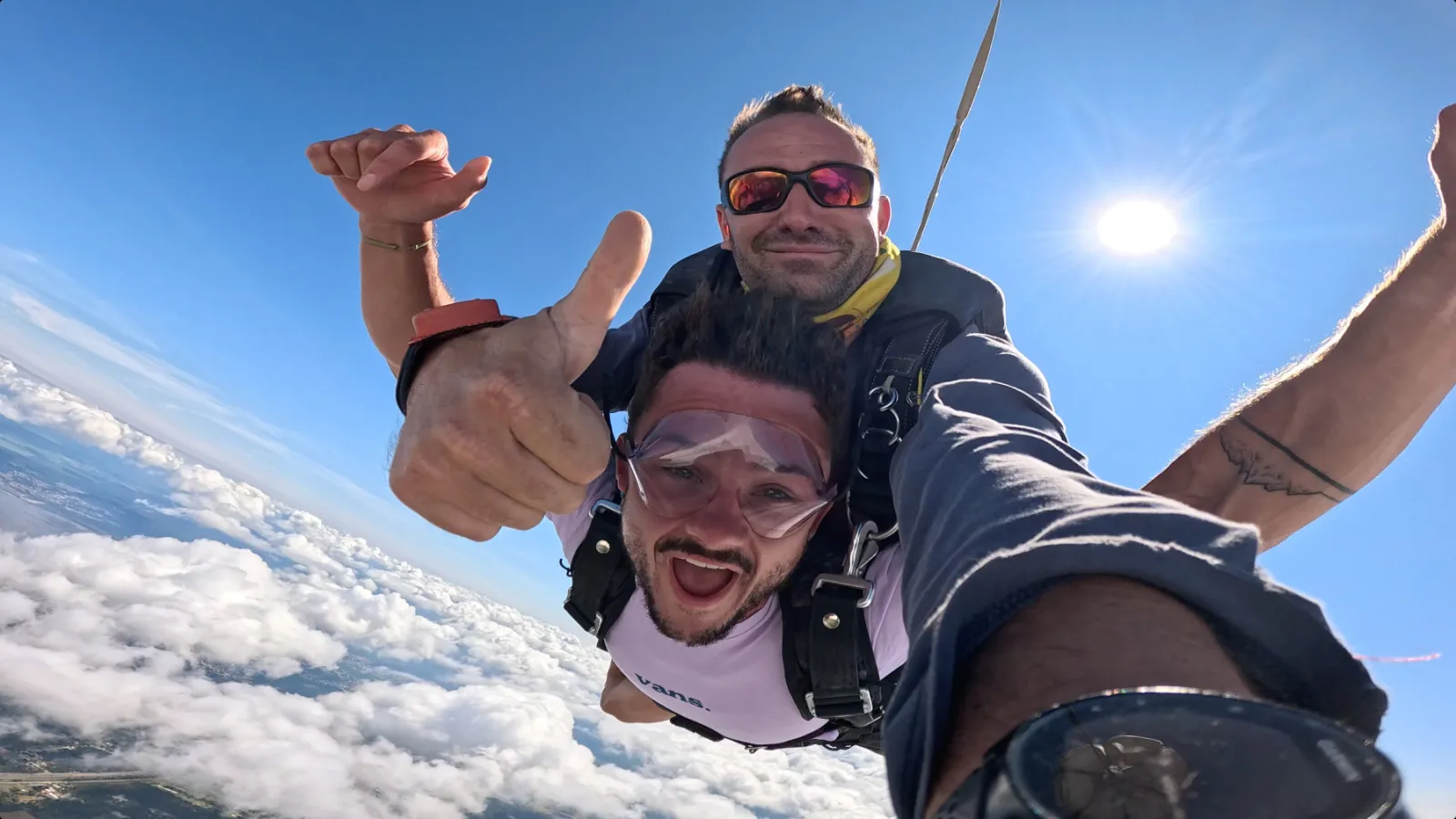 Two men skydiving in tandem smile at the camera while flying above the clouds with blue sky in the background during a parachute jump over Bordeaux.