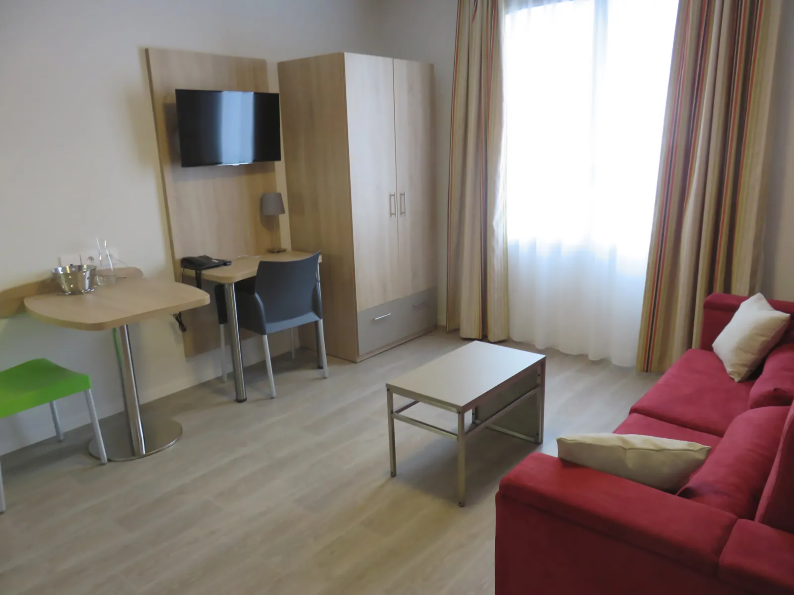 Spacious hotel suite featuring a red sofa, desk with flat-screen TV, wardrobe storage, coffee table and access to a balcony overlooking Bordeaux