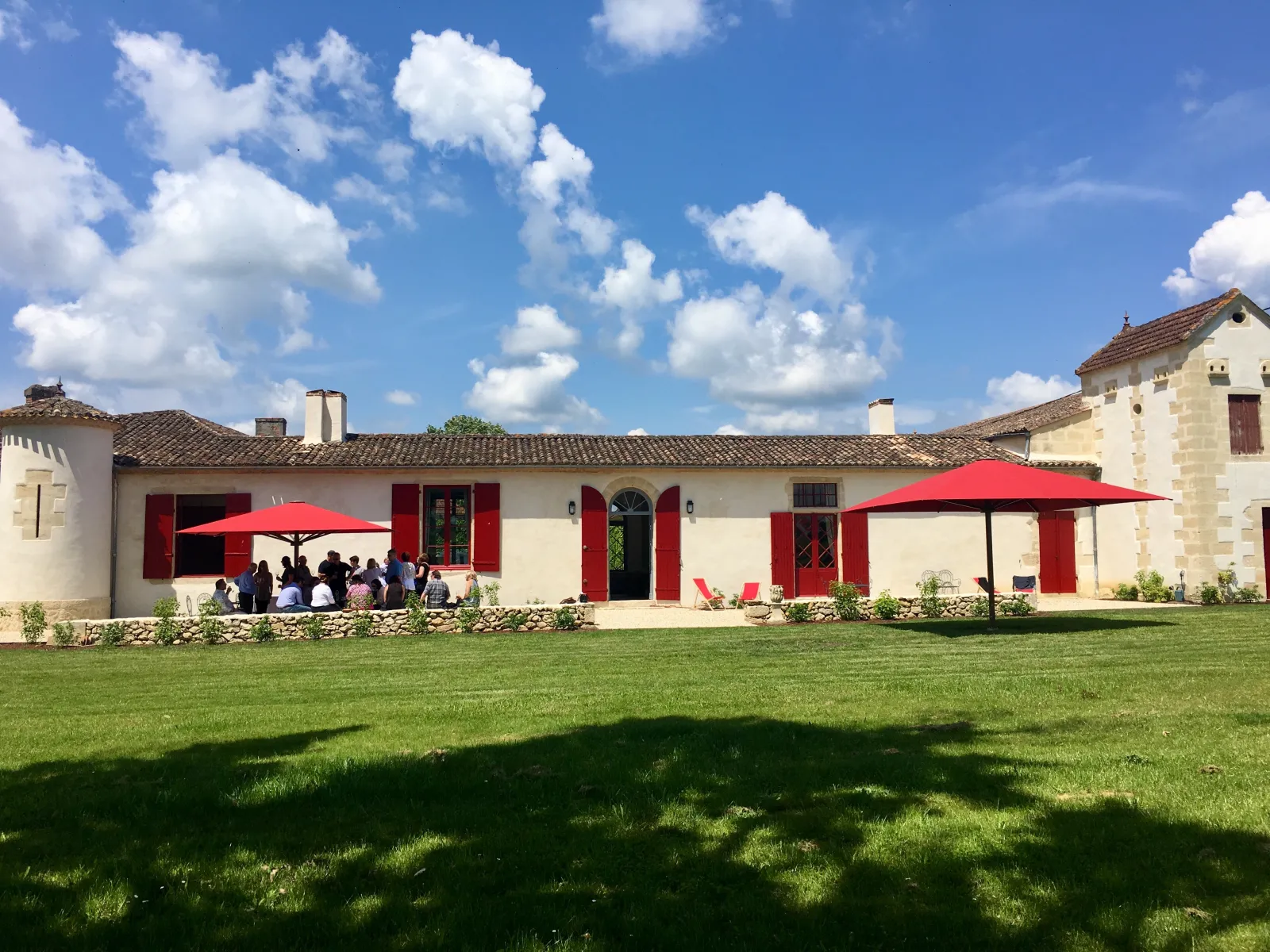 Sunny view of Château Sigalas Rabaud in Bordeaux featuring white facades with red shutters, green lawn in foreground, red umbrellas and visitors gathered under canopies, blue cloudy sky.