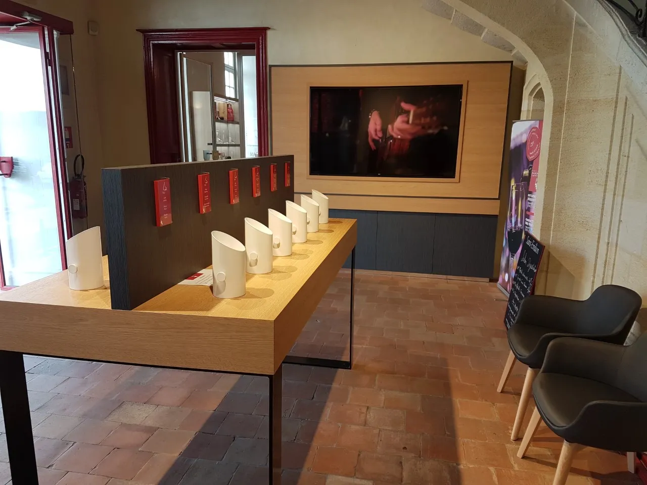 Interior of the Maison du Vin de Saint-Emilion wine museum in Bordeaux, featuring an interactive exhibition with illuminated displays, black chairs, wall-mounted screen, and wine presentation elements.