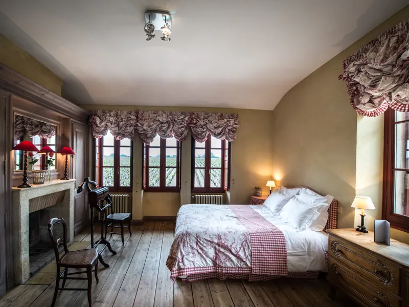 Elegant bedroom at Château Giscours featuring a bed with red and white checkered bedding, ornate fireplace, panoramic windows overlooking the vineyard, patterned curtains and classic furniture