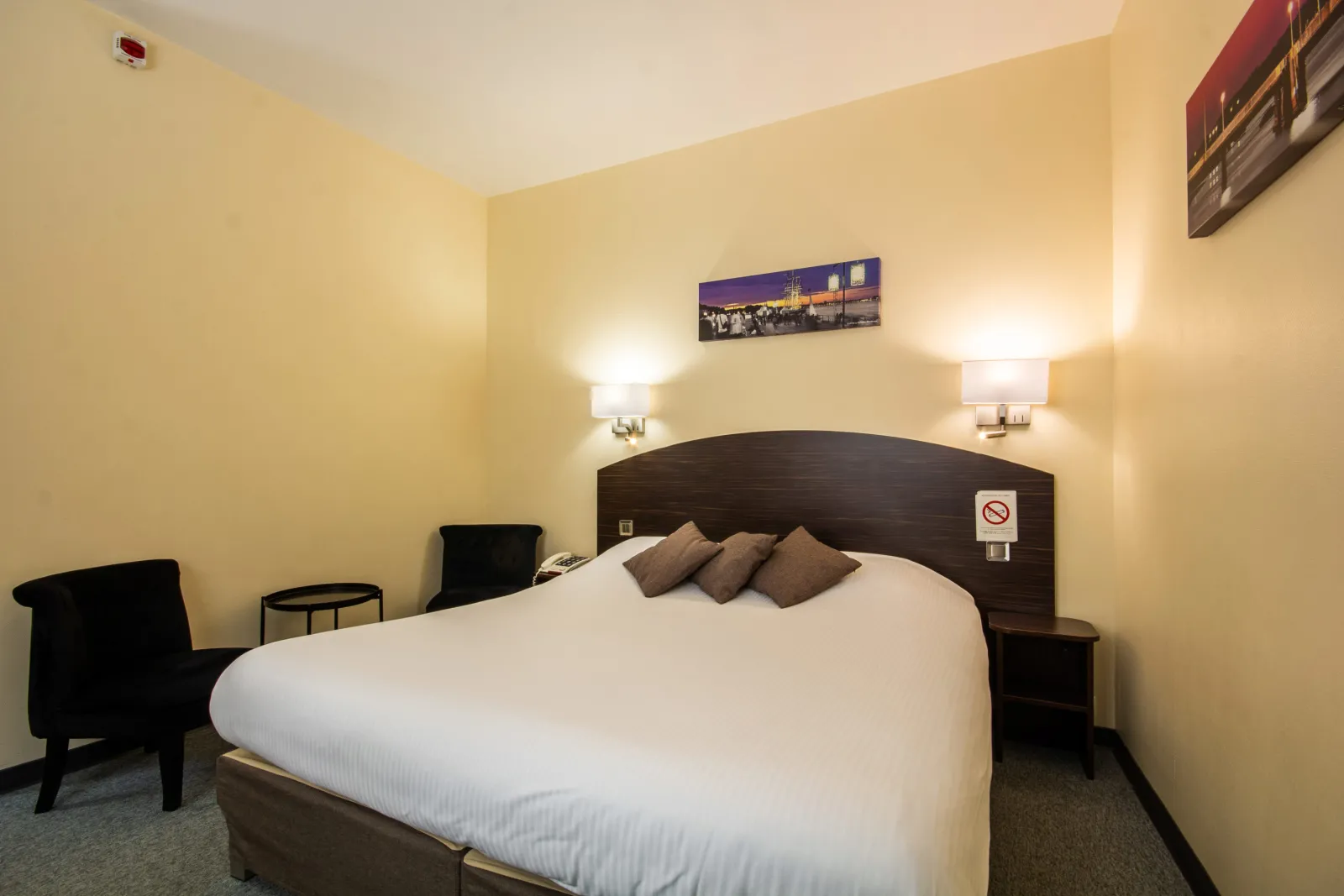 Modern hotel bedroom featuring a double bed with white linens, dark wooden headboard, pale yellow walls, wall-mounted lamps, black armchairs, and a Bordeaux cityscape photograph above the bed