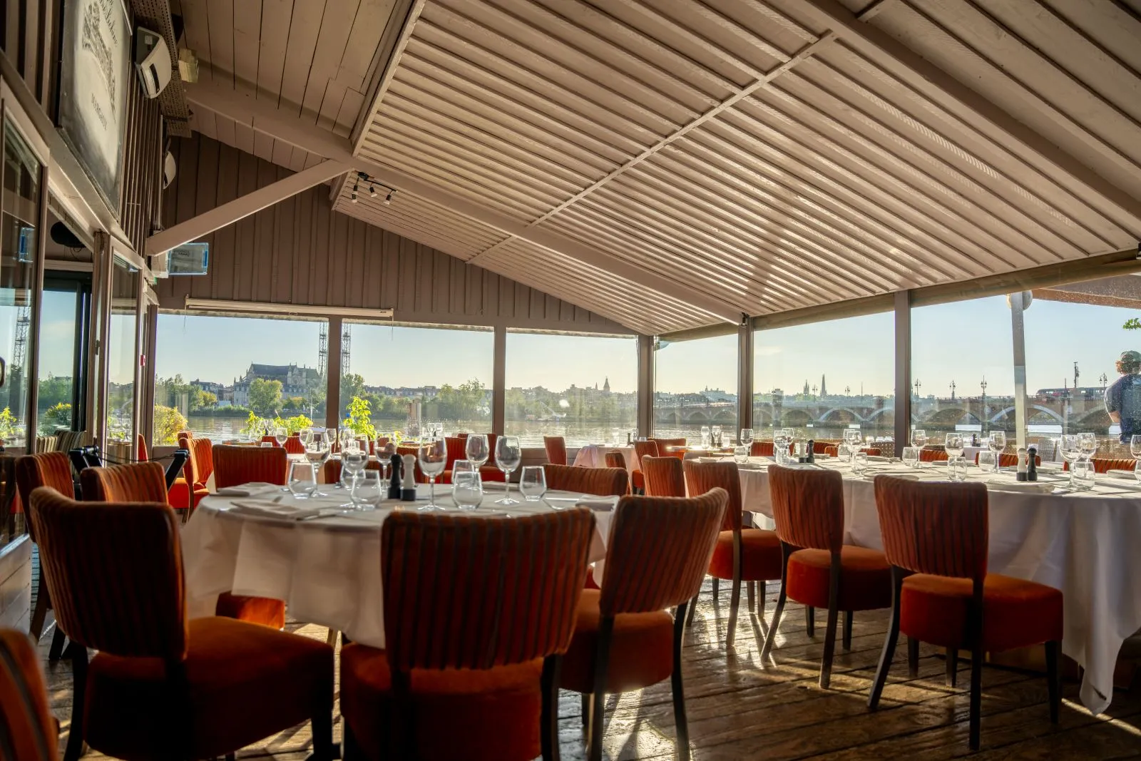 Panoramic restaurant dining room with round tables, orange chairs, and floor-to-ceiling windows overlooking the Garonne River and Bordeaux cityscape.