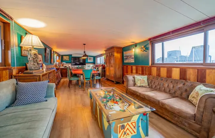 Spacious and elegant interior of a converted riverboat in Bordeaux, featuring warm wooden tones, tufted sofa, colorful coffee table, equipped kitchen in the background, and large windows overlooking the city and river.