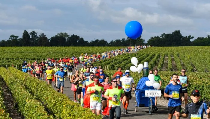 A large group of marathon runners participating in the Châteaux du Médoc Marathon running through a Bordeaux vineyard with a giant blue balloon and support signs.