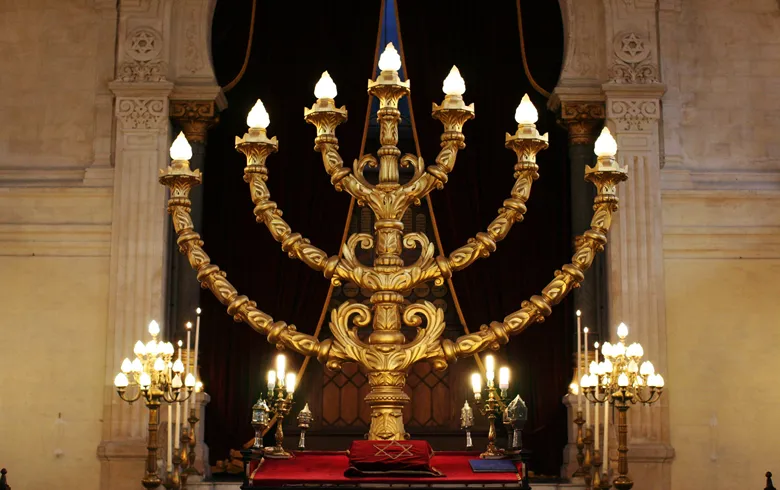 Golden seven-branched menorah lit in the Bordeaux Synagogue, with side candelabras and classical architecture in the background