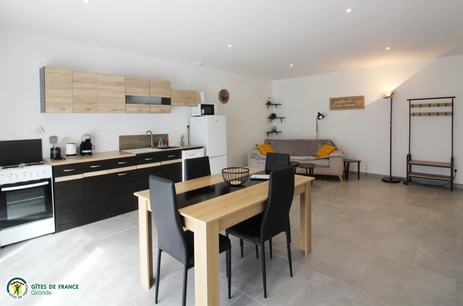Spacious interior of the Tanins guesthouse at Château Pierre de Montignac in Bordeaux, featuring an equipped kitchen, dining area with wooden table, and gray sofa with yellow cushions in a modern, bright open-plan space.