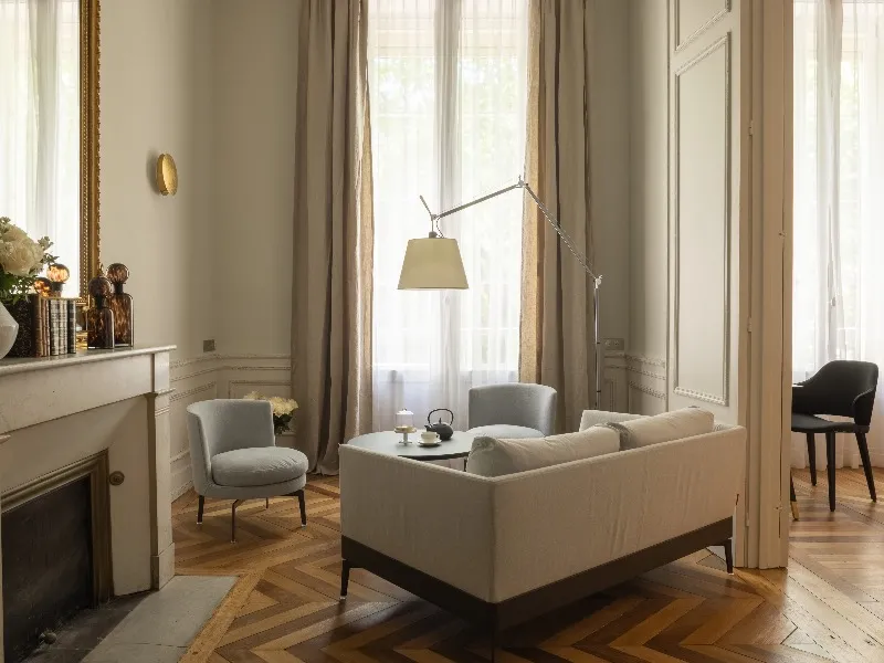 Elegant living room at Hôtel Villas Foch in Bordeaux featuring a beige sofa, gray armchairs, herringbone parquet flooring, high ceilings with moldings, tall draped windows, designer floor lamp and classic fireplace