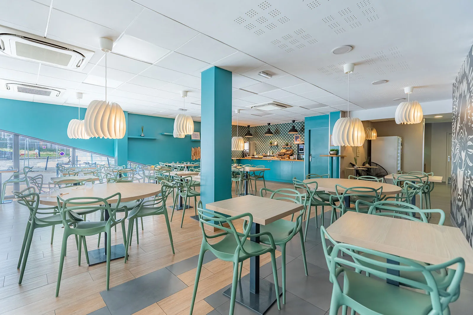 Modern common room in an aparthotel featuring turquoise walls, beige tables, green designer chairs, white pendant lights, and equipped kitchen in the background.