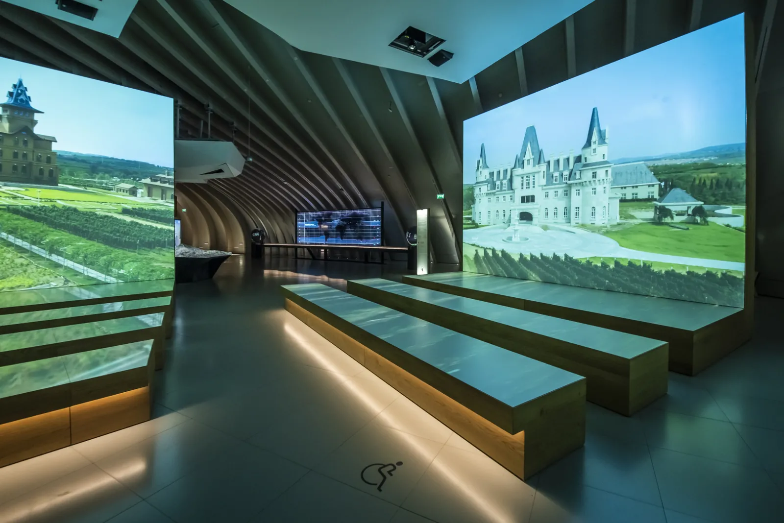 Modern interior of Cité du Vin in Bordeaux featuring large projection screens displaying wine châteaux, curved wooden ceiling architecture, polished concrete floor, and illuminated seating.
