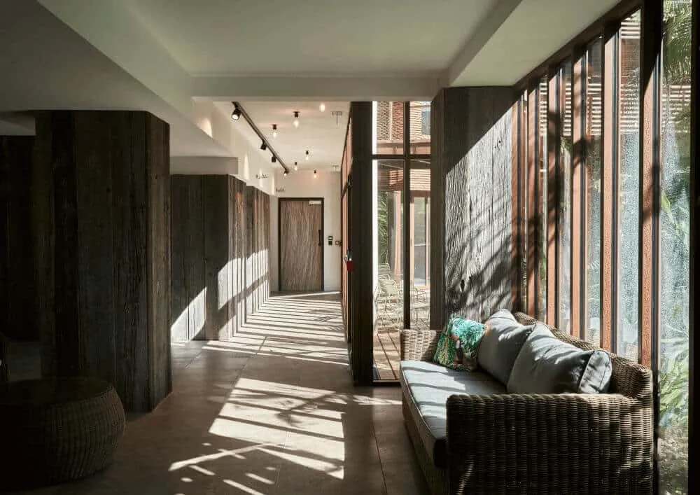 Modern corridor at La Zoologie spa in Bordeaux featuring dark wooden doors, large windows with natural light, woven seating bench, and views of exterior greenery.