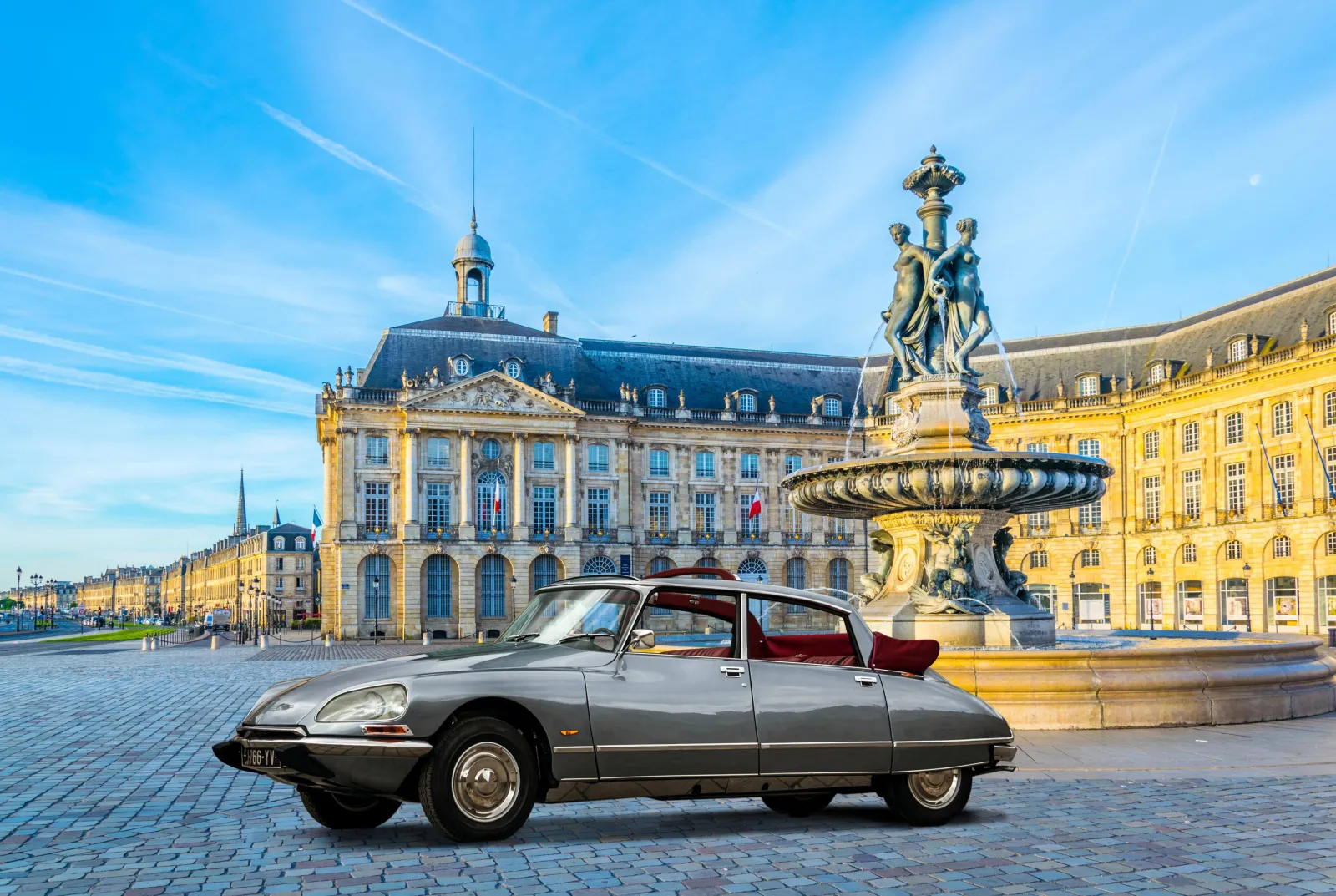A vintage grey Citroën DS with red interior parked at Place de la Bourse in Bordeaux, featuring the Three Graces fountain and historic buildings in the background