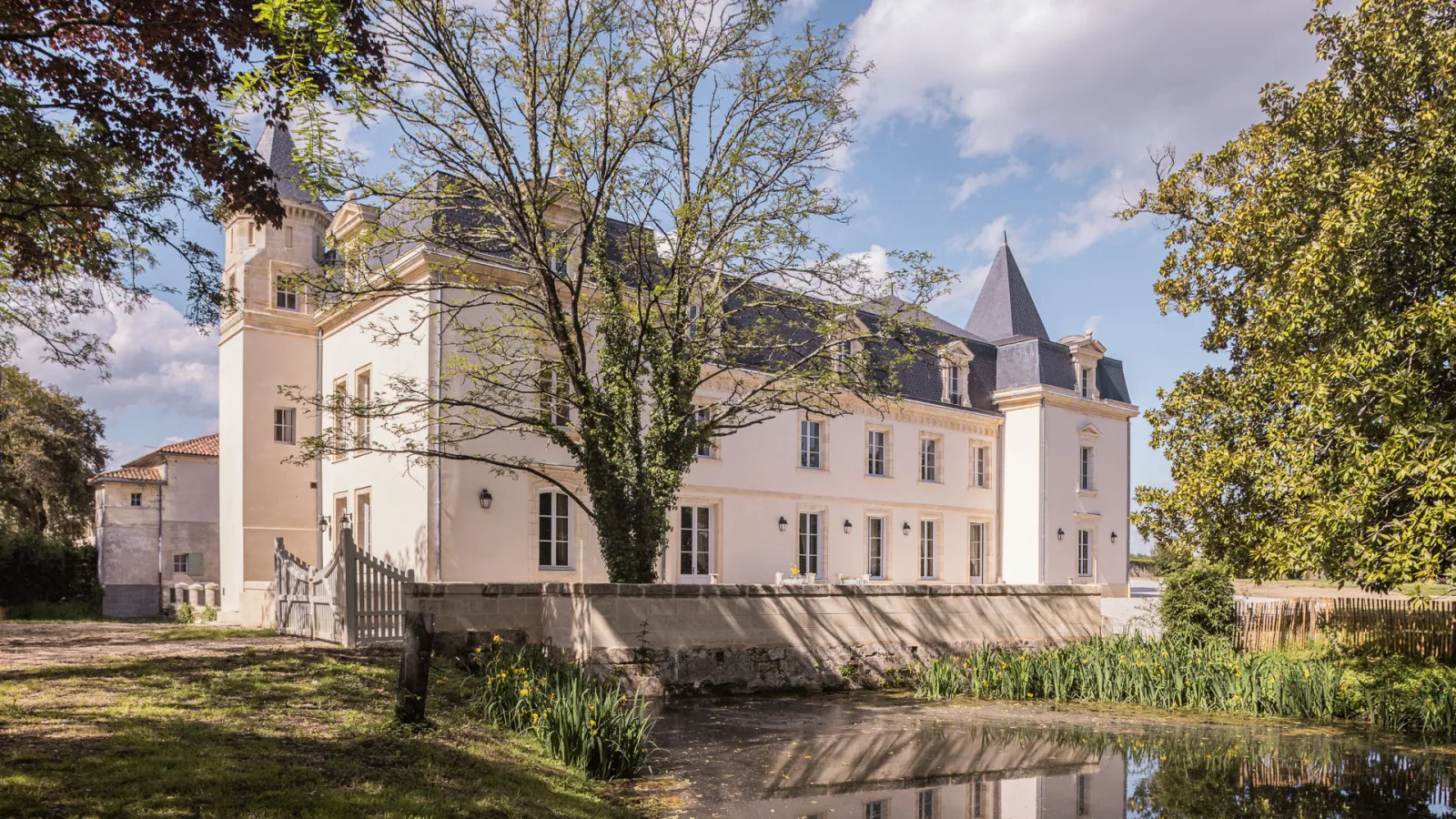 Sénéjac Castle near Bordeaux, an elegant 18th-century mansion featuring distinctive turrets, pale pink and white facade, surrounded by park grounds with a water channel and trees.