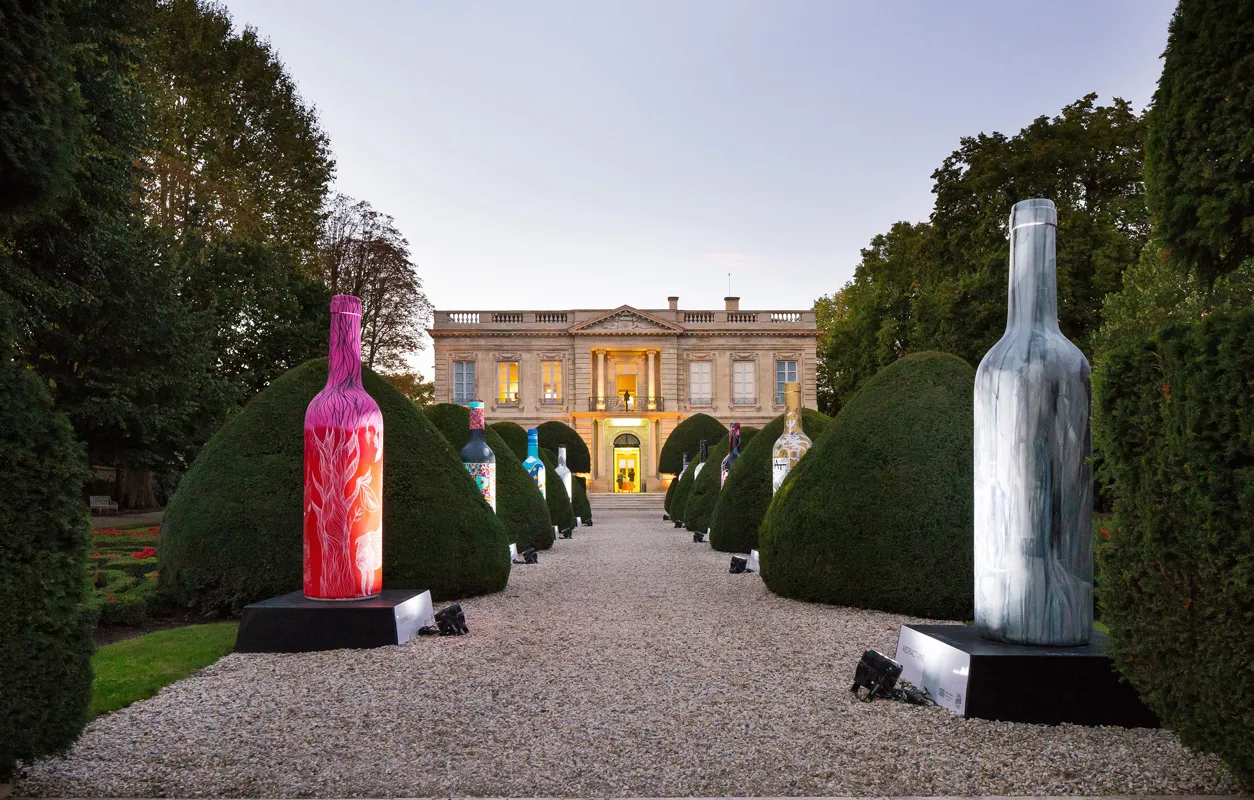 Giant oversized wine bottles displayed as art installations in a French formal garden with trimmed hedges leading to an illuminated neoclassical mansion