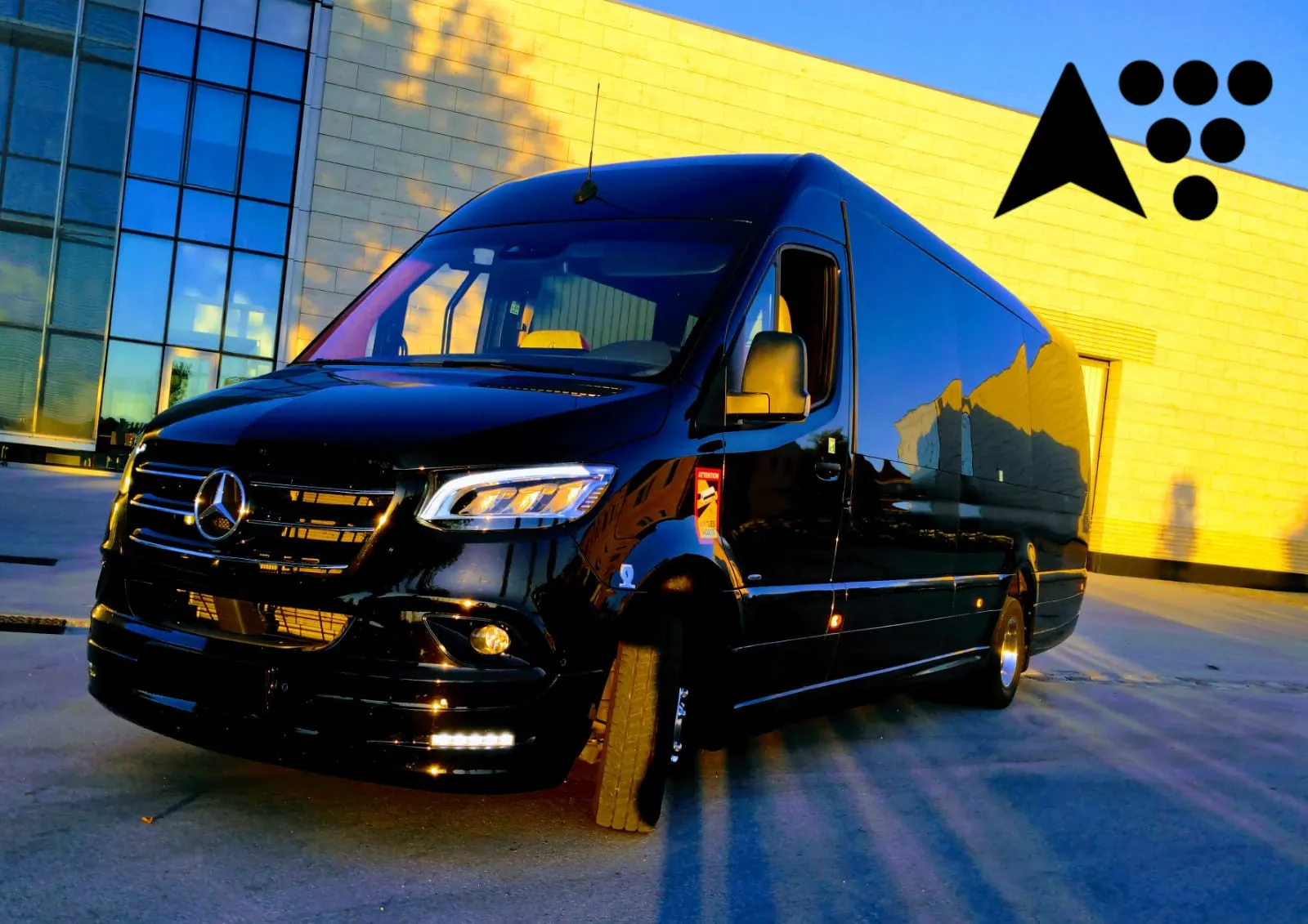 A black Mercedes minibus from BTA (Bordeaux Tourisme Affaires) parked in front of a modern building with yellow facade and blue windows, photographed in late afternoon with headlights on.