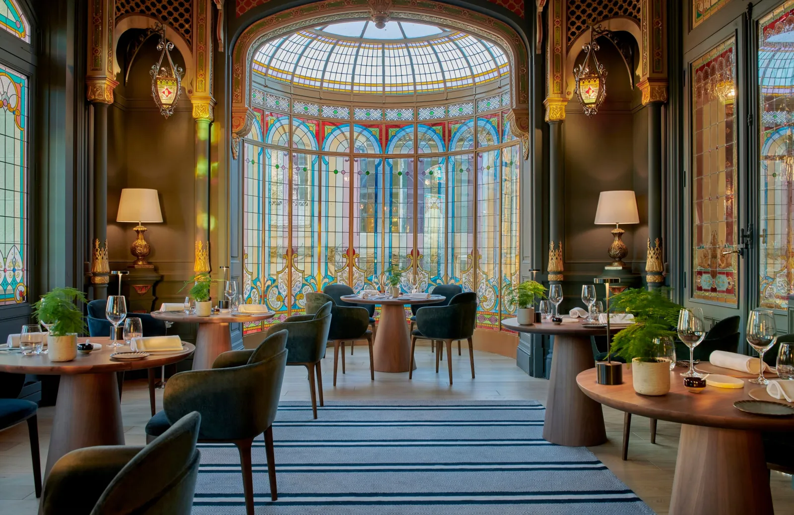 Elegant restaurant dining room featuring ornamental glass windows, round wooden tables, blue chairs, golden wall lamps, and a geometric glass dome ceiling with colorful panes.