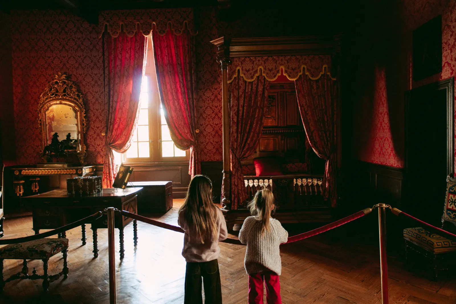 Two children viewing an ornately decorated royal bedroom at Château Royal de Cazeneuve, featuring red damask wallpaper, crimson drapes, a four-poster canopy bed, and natural light from a tall window.