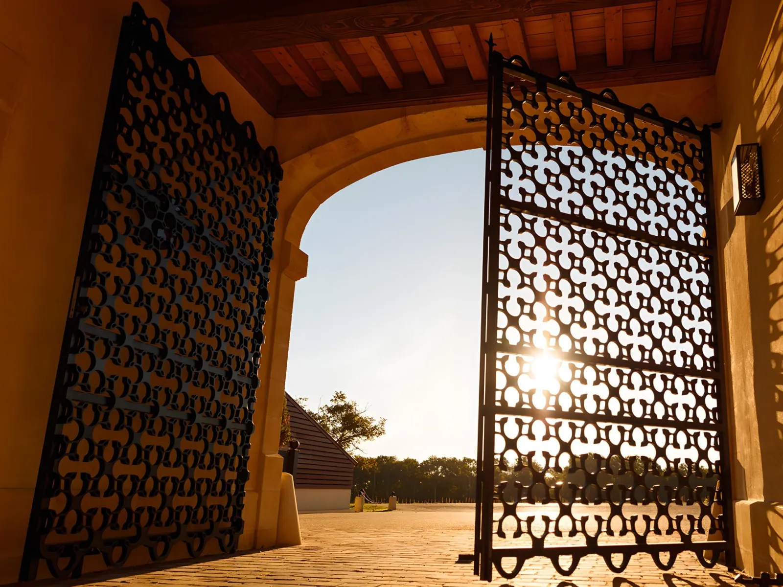 View through two ornate wrought iron gates with geometric cutout patterns framing a sunlit courtyard with historic building and trees at Château du Lort in Bordeaux