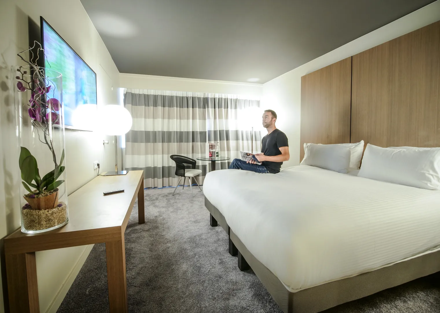 Modern hotel room at Pullman Bordeaux Lac featuring a man sitting in a chair, a large white bed, a desk with orchid, flat-screen TV, and gray and white striped curtains.