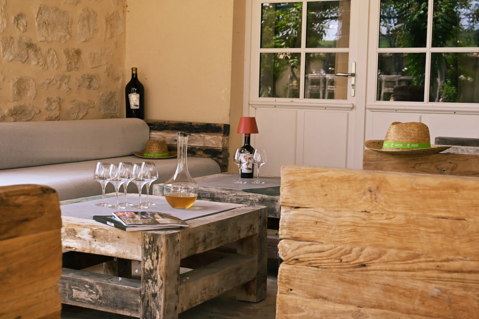 Elegant lounge at Domaine des Perchés in Bordeaux featuring raw wood rustic furniture, wine glasses, wine bottle, red lamp, straw hats, and glass doors overlooking a green garden