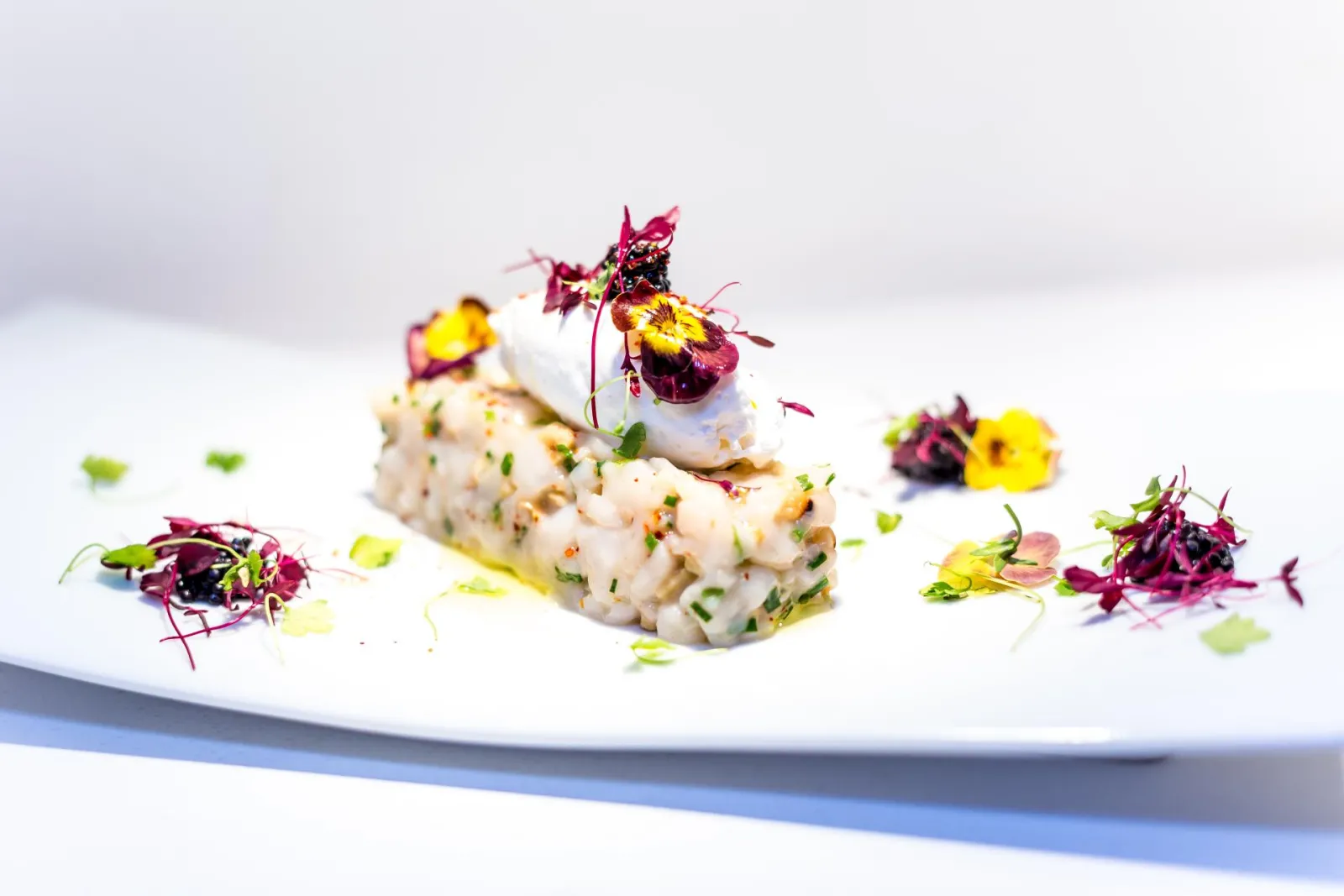 Fine dining plate featuring fish tartare topped with white foam, fresh herbs, and purple and yellow edible flowers, presented on white with minimalist plating.
