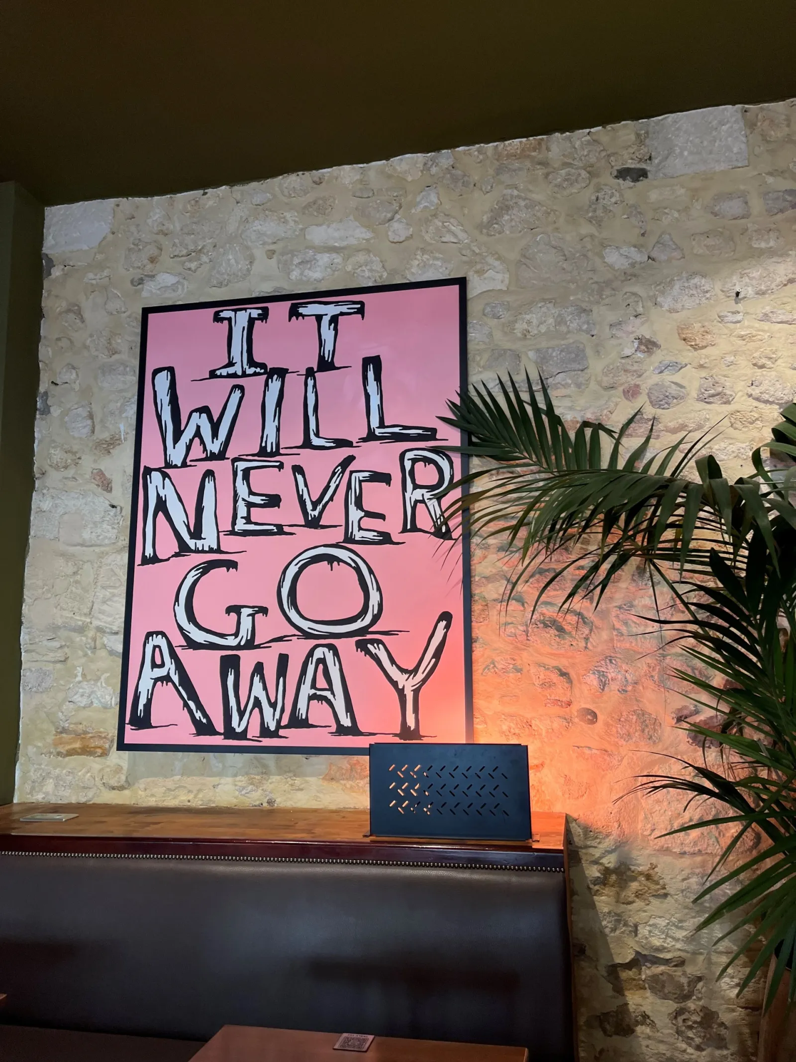 Pink poster with black text reading 'IT WILL NEVER GO AWAY' on a stone wall with a green plant, at Cajou Caffé in Bordeaux