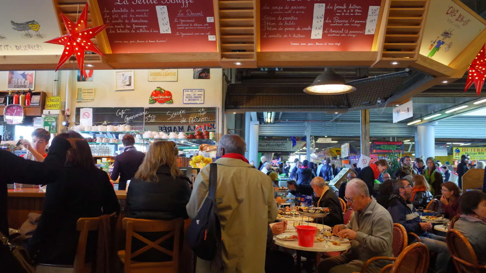 Interior view of Marché des Capucins in Bordeaux showing seated customers at tables, a service counter in the background, illuminated red menu boards, and Christmas decorations including a large lit red star.