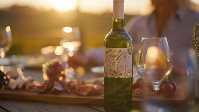 Bordeaux white wine bottle on an outdoor tasting table at sunset, with empty glass and guests in the background