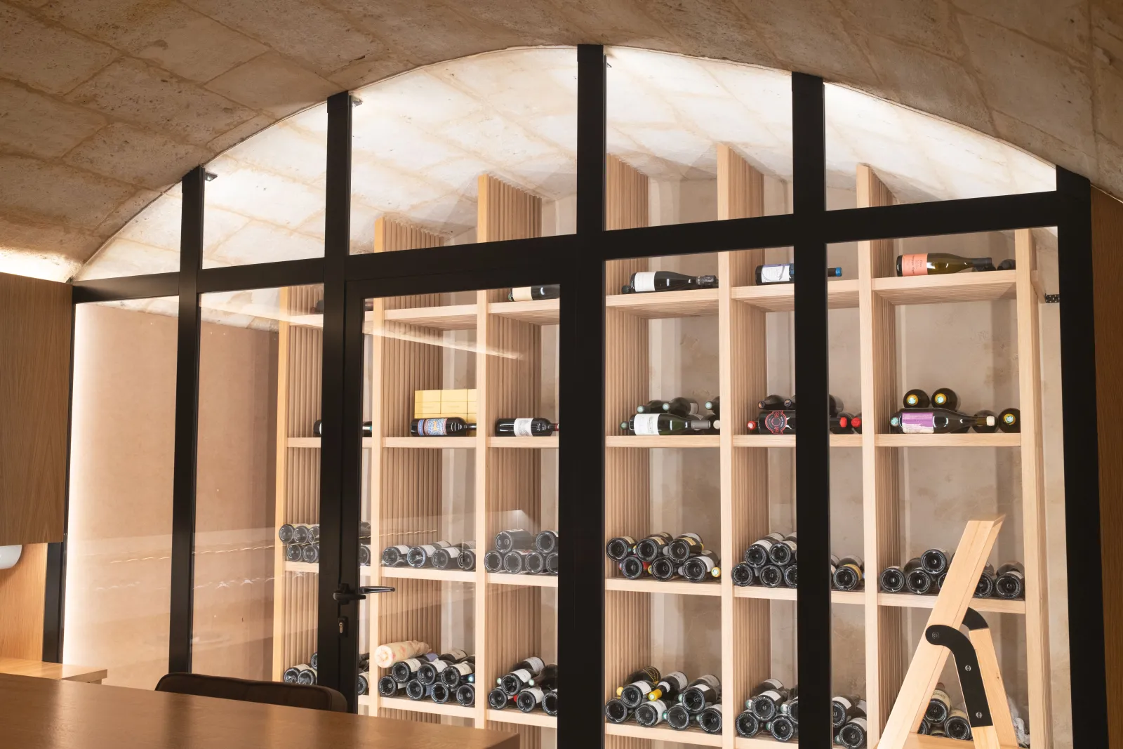 Wine cellar display cabinet with black frame and glass doors, showcasing wine bottles and accessories on light wood shelves in Bordeaux.