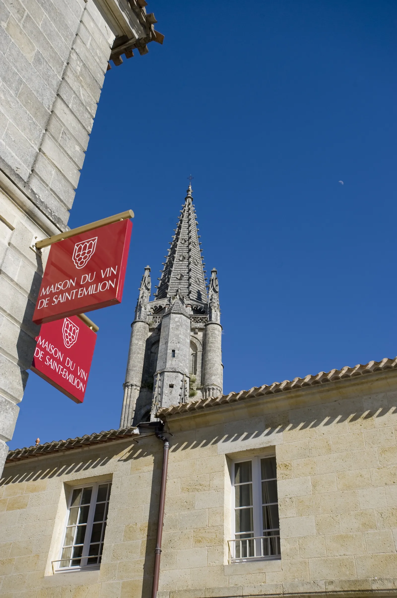 Facade of the Maison du Vin de Saint-Emilion in Bordeaux with red flags and gothic bell tower in the background under blue sky
