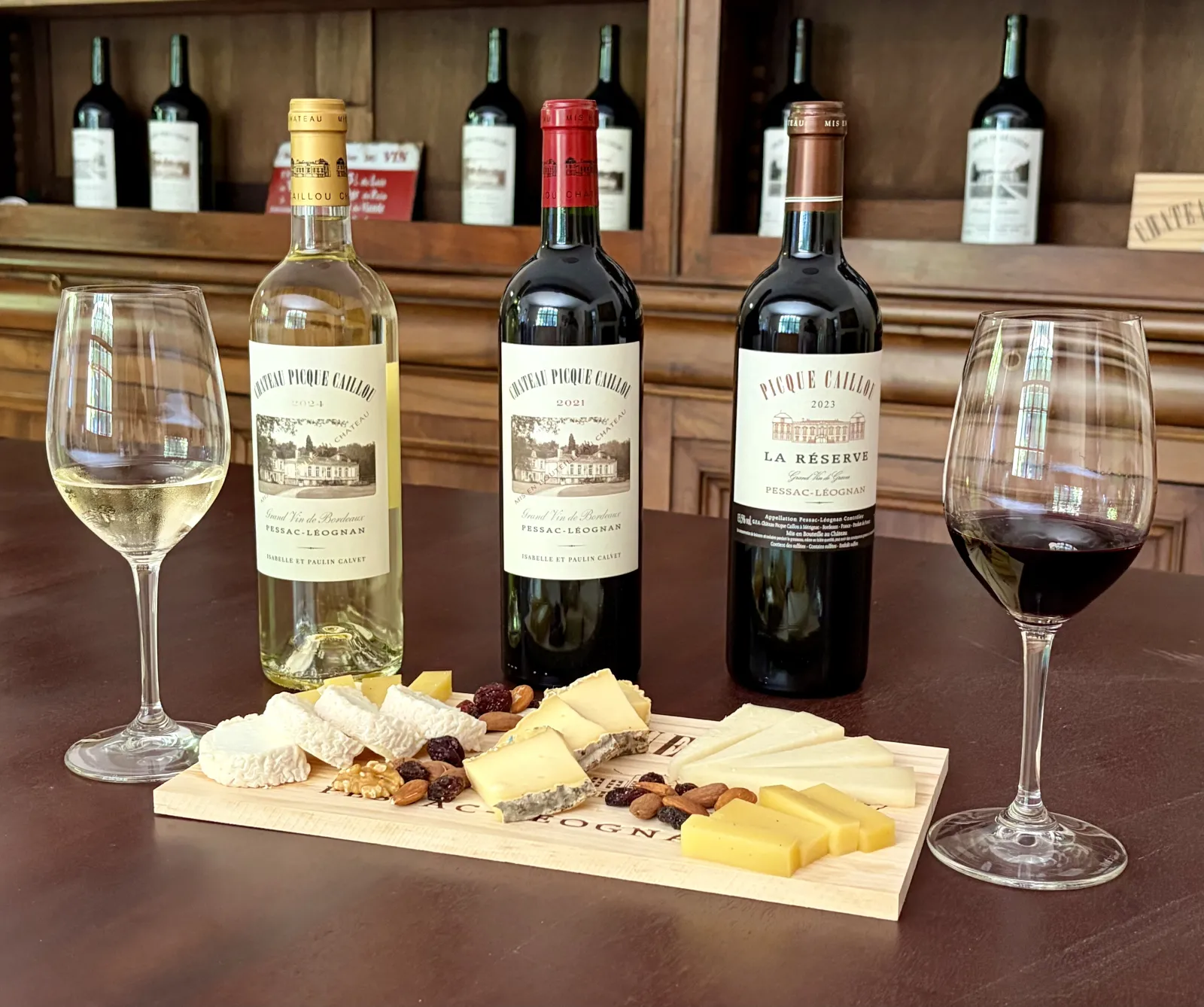 Wine tasting from Château Picque Caillou in Bordeaux featuring three wine bottles, two filled glasses, and a wooden board with cheese and dried fruits against a cellar background.
