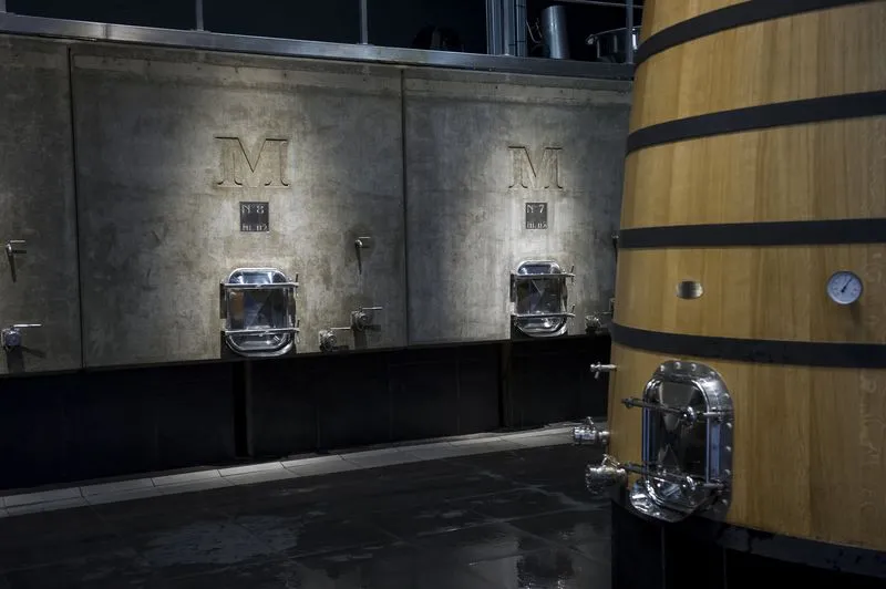 Modern winery interior featuring two metal fermentation tanks marked with an M, a large oak barrel with black bands, and professional winemaking equipment at Château Malescasse in Bordeaux.