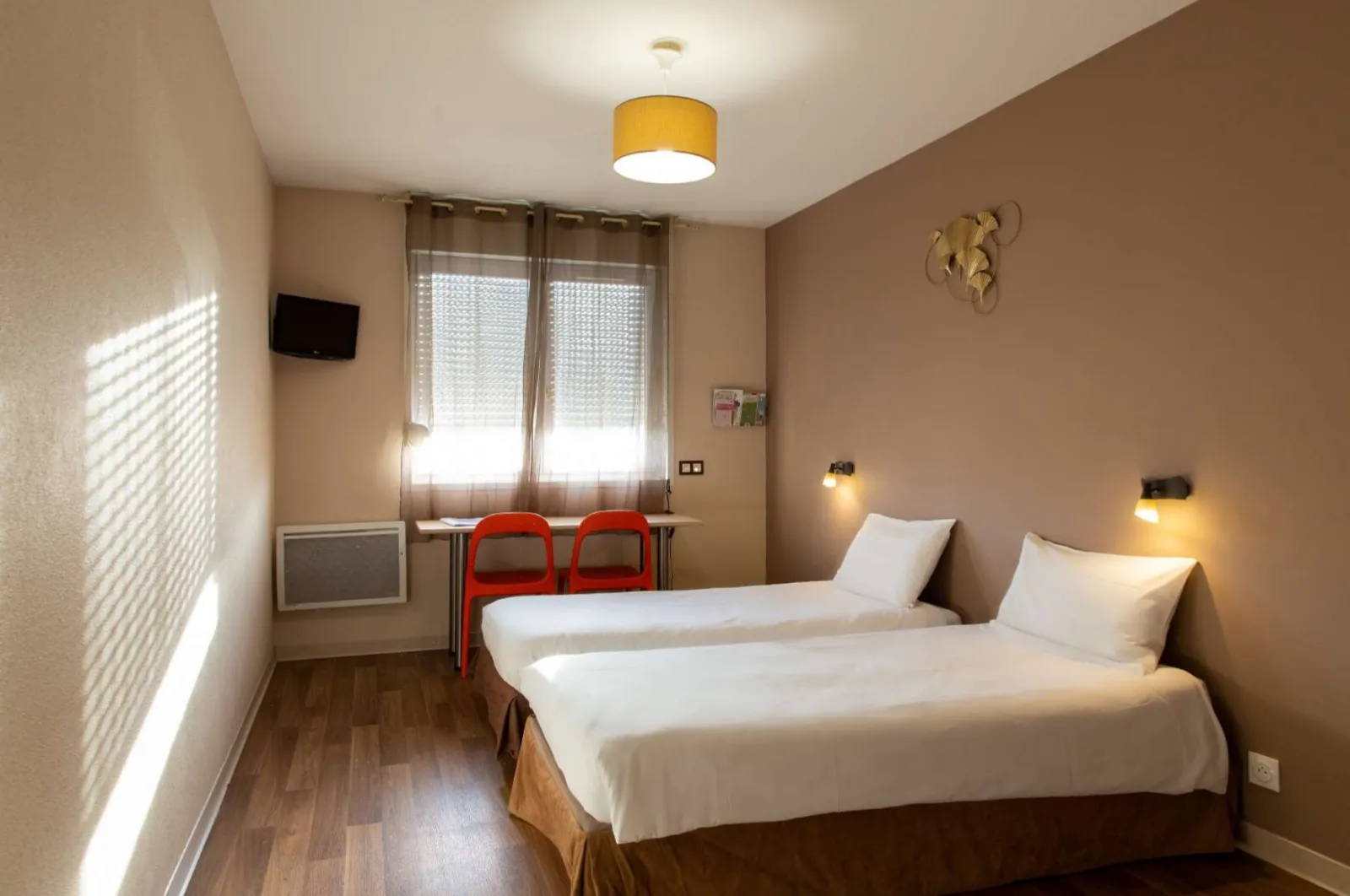 Youth hostel double room with two single beds with white linens, red chairs, window with blinds, beige walls and modern lighting fixtures