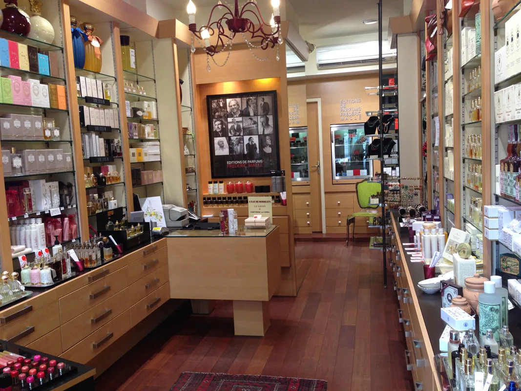 Elegant interior of a perfume shop featuring a central counter, light wood shelves displaying fragrance bottles, decorative chandelier, and artist photographs on the wall.